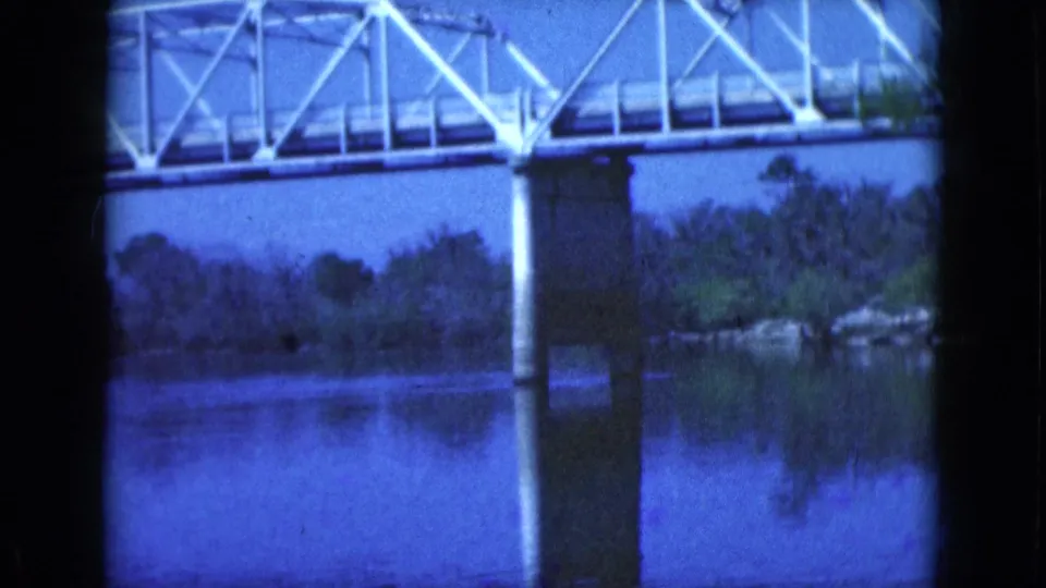 1975 Florida 8mm frame showing a white truss bridge, concrete pier, river reflection, and tree-lined bank.