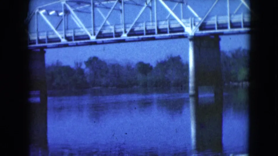 Final 1975 Florida 8mm frame of a white truss bridge, vertical pier, reflected water, and trees across the river.