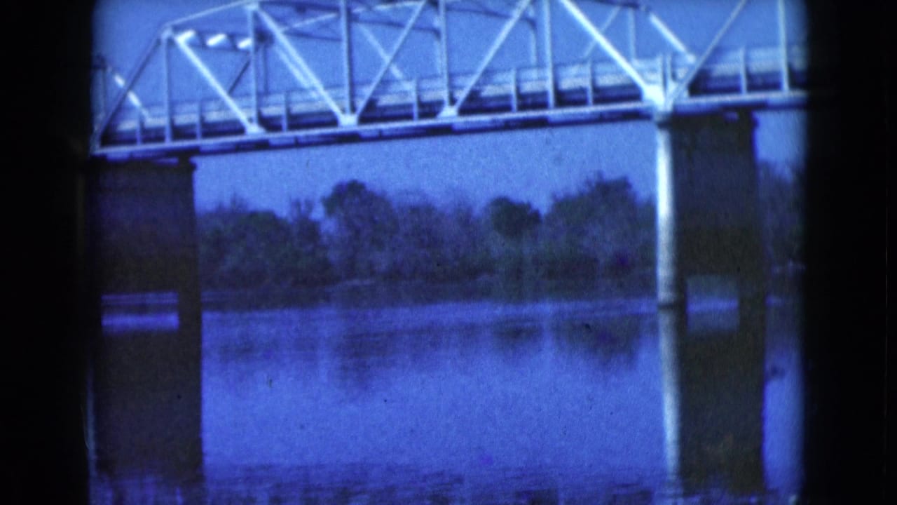 1975 Florida 8mm frame showing a pale metal truss bridge over a calm river, with concrete piers, tree line, reflections, and blue film tint.