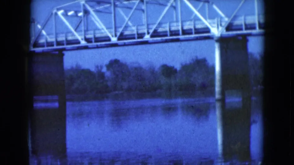 1975 Florida 8mm frame showing a pale metal truss bridge over a calm river, with concrete piers, tree line, reflections, and blue film tint.