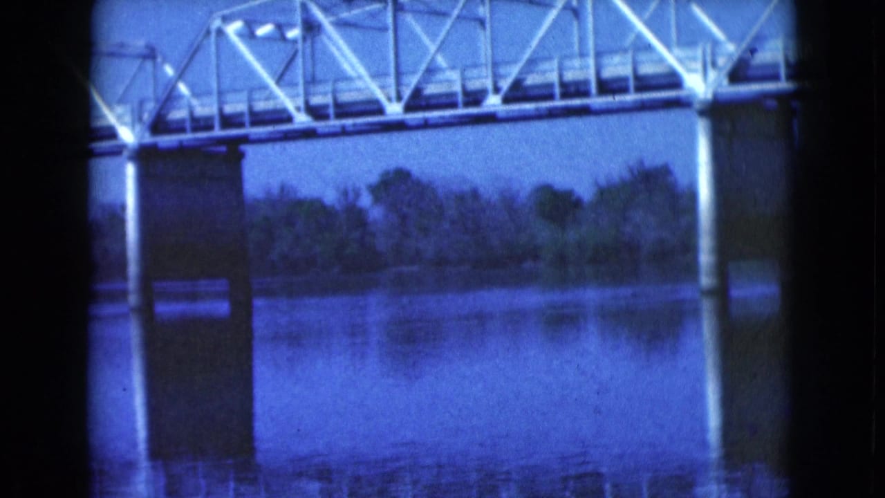 1975 Florida home-movie frame of a metal bridge span and central concrete support reflected in still water, with trees along the far shoreline.