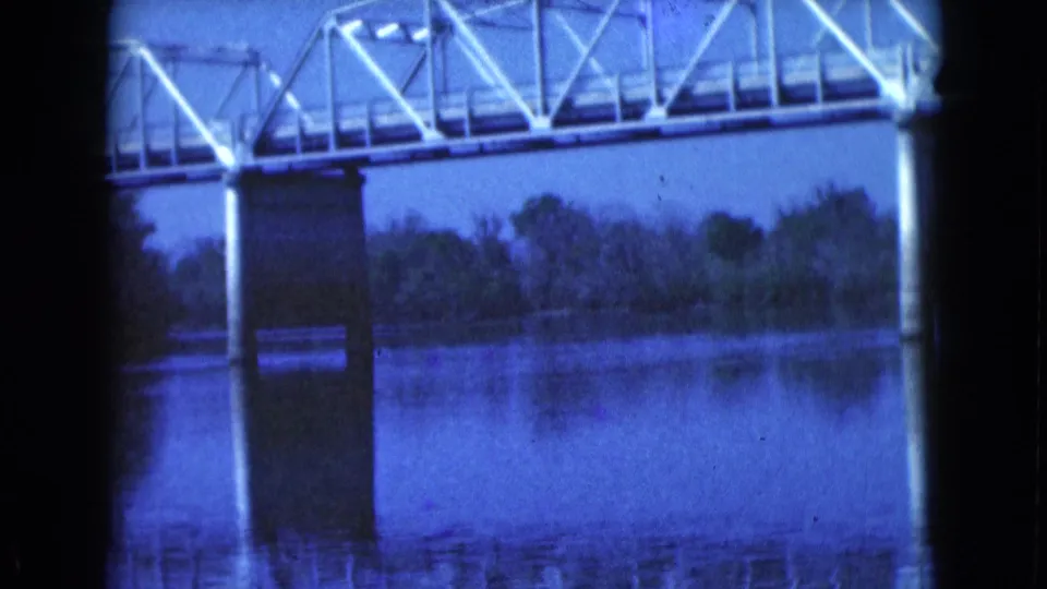 Blue-tinted 8mm frame from 1975 Florida showing the truss bridge edge, concrete supports, calm water, and a tree-lined opposite bank.