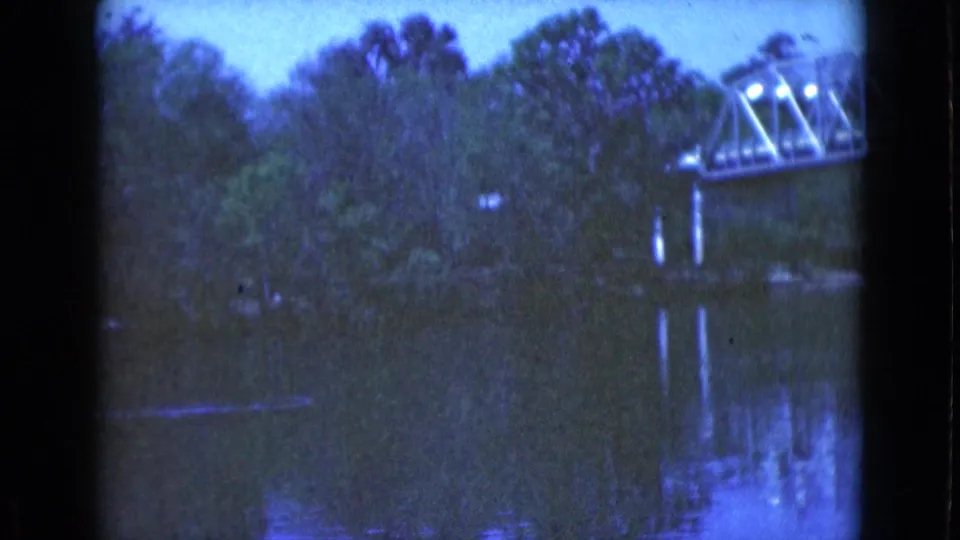 1975 Florida 8mm frame with the far end of a metal truss bridge at right, dense trees, dark calm water, and vertical bridge reflections.