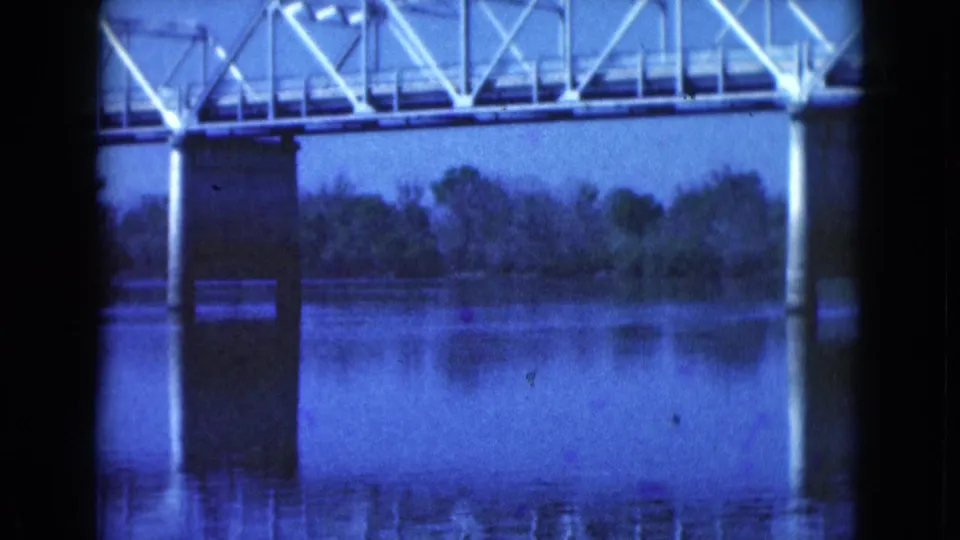 1975 Florida home-movie frame of a white truss bridge and support pier reflected in a still river.