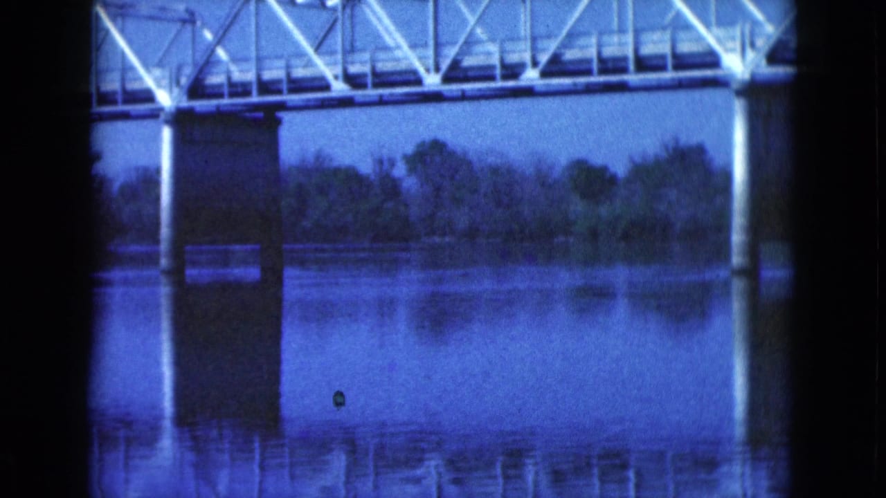 Metal truss bridge crossing a calm lake with reflected supports.