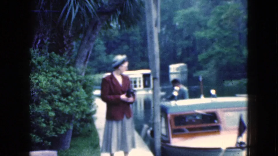 A woman in a brown jacket and skirt stands near a small boat amid greenery in 1975 Florida home-movie footage.