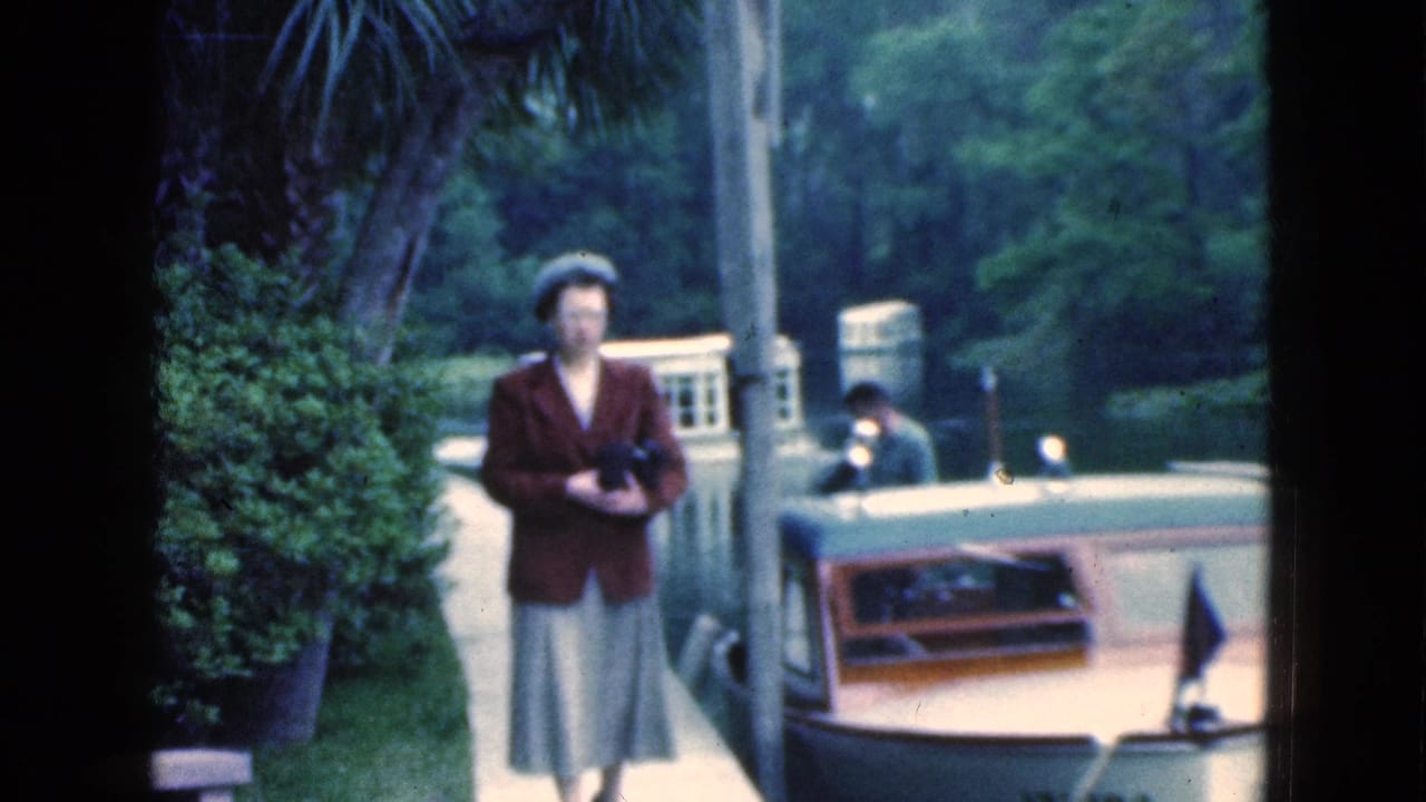 In 1975 Florida home-movie footage, a woman in a maroon jacket holds a camera beside a canal while boats move nearby.