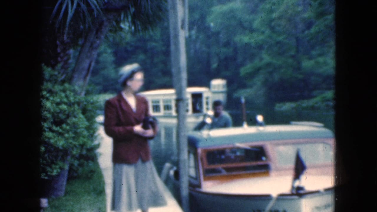 A woman holding a camera looks along a Florida canal, with a small boat, larger cabin vessel, and dense greenery behind her.