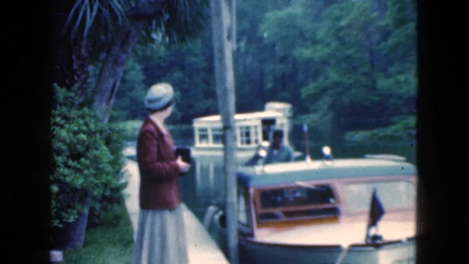The woman walks along a Florida canal path in 1975, holding a camera while looking toward boats moving through the waterway.
