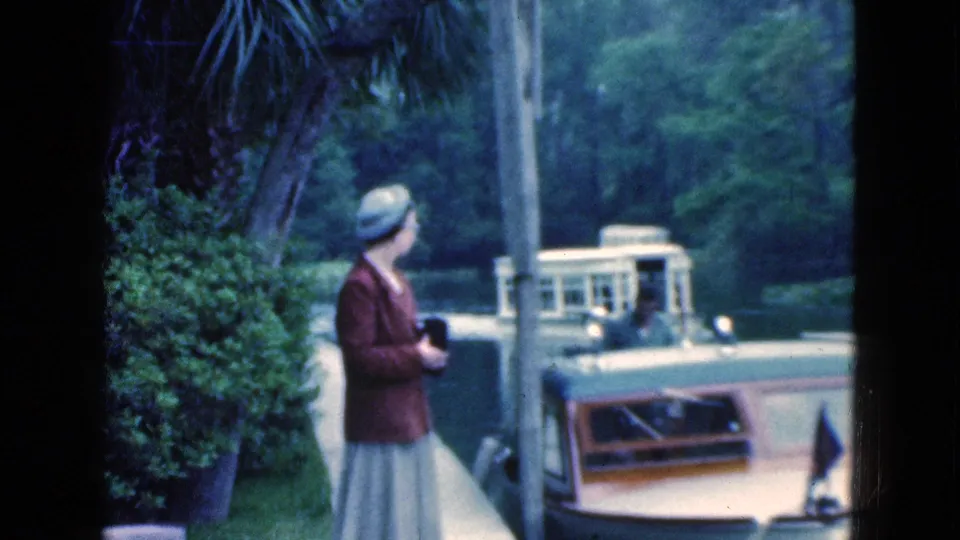 A profile view shows the woman with a camera beside a Florida canal, watching a boat pass against a tree-lined background.