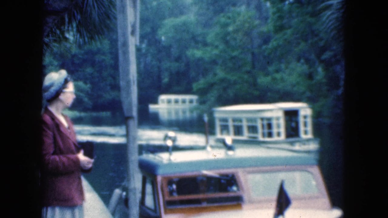 Woman in a brown coat stands on a dock near boats on a wooded waterway.