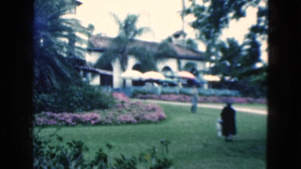 1975 Florida 8mm frame showing landscaped resort-style grounds with palms, flower beds, umbrellas, an arched building, and two distant people walking.