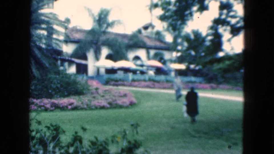 1975 Florida 8mm frame of people walking through resort-style grounds with palms, flower beds, umbrellas, lawn, and an arched building.