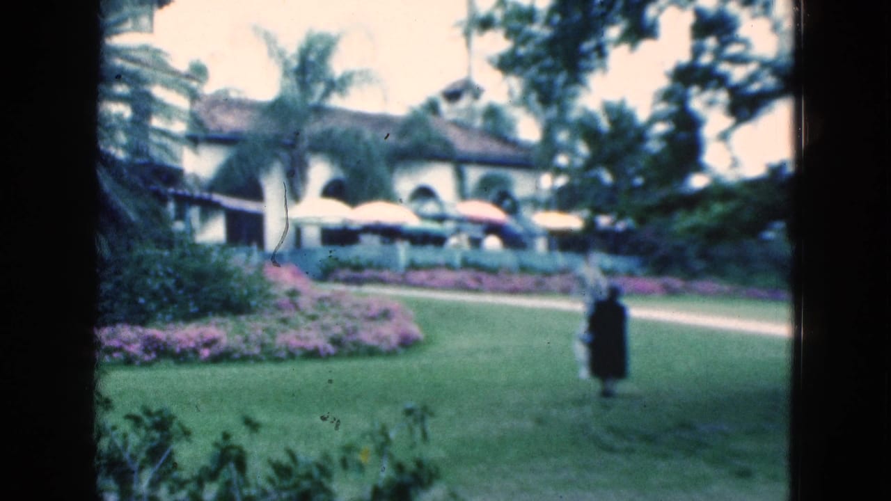 Adults walk across a green lawn near flower beds and a white arched building.