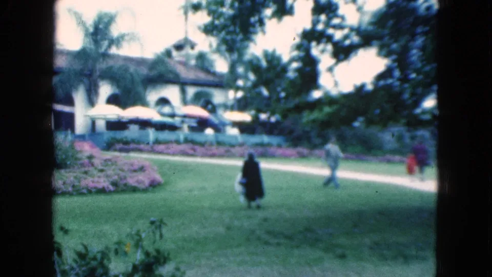 Soft 8mm view of 1975 Florida garden grounds with adults walking near pink beds, palms, and a Spanish-style building.