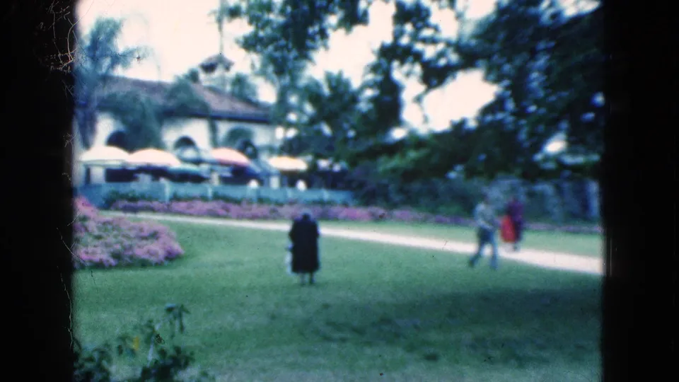 1975 Florida frame showing distant adults on landscaped grounds with flower beds, palms, umbrellas, and a white arched building.