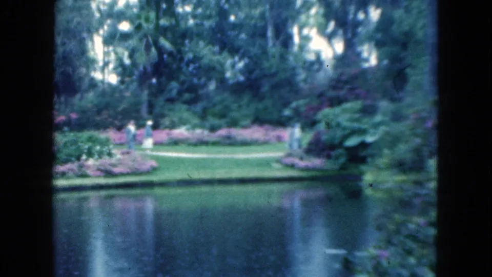 Soft 1975 Florida 8mm view of people walking along a curved garden path beyond a reflective lake.