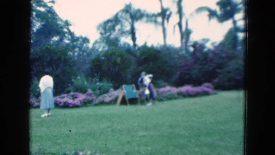 1975 Florida home-movie frame with two women crossing a landscaped backyard near a folding chair, flowers, lawn, and palm trees.