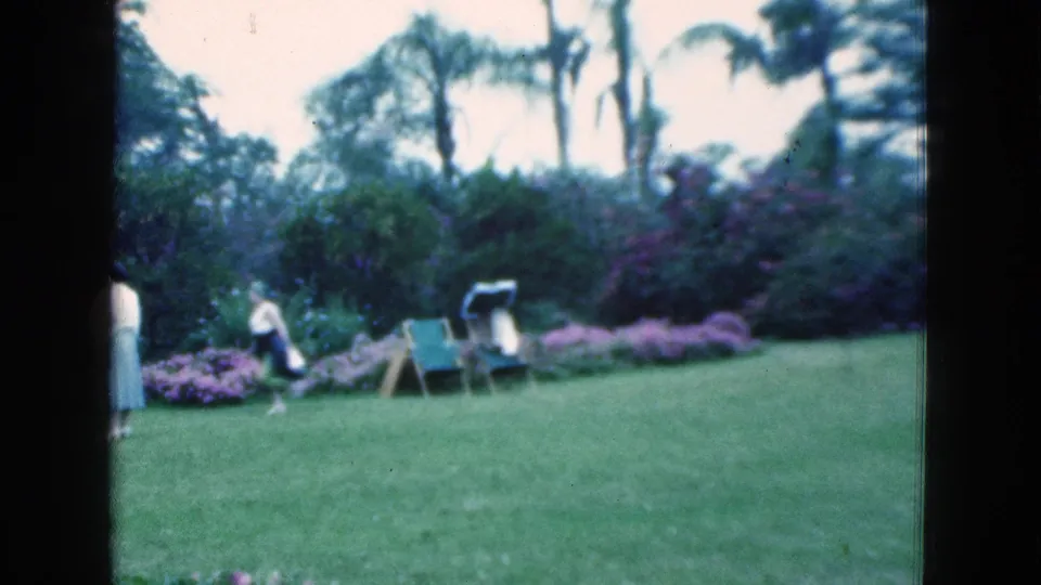 Soft 8mm view of two women in a 1975 Florida backyard garden, with a folding chair, flowering plants, lawn, and palms visible.