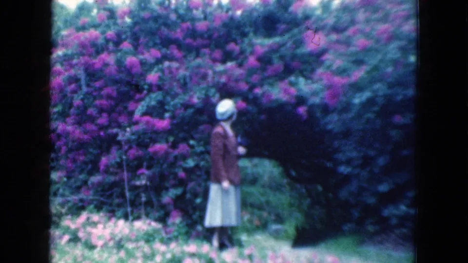 A woman in a brown jacket and light skirt stands near flowering bushes in a 1975 Florida garden scene.