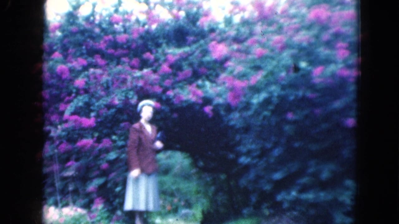 In 1975 Florida home-movie footage, a woman in a brown jacket and pale skirt stands near dense flowering shrubs and greenery.