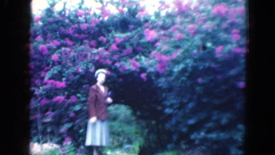 In 1975 Florida home-movie footage, a woman in a brown jacket and pale skirt stands near dense flowering shrubs and greenery.