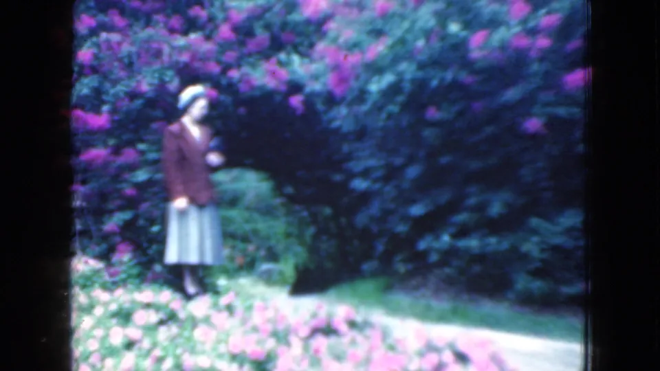 A woman in a brown jacket stands near a curved garden path, with pale and purple flowers visible in the foreground.