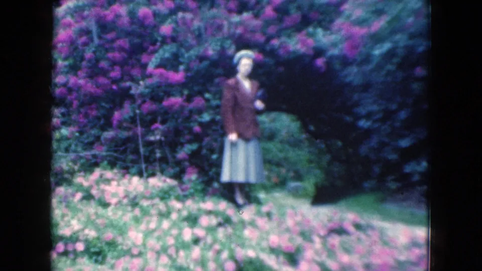 The woman remains beside thick flowering shrubs and a narrow path, surrounded by saturated greens and pink-purple blossoms.