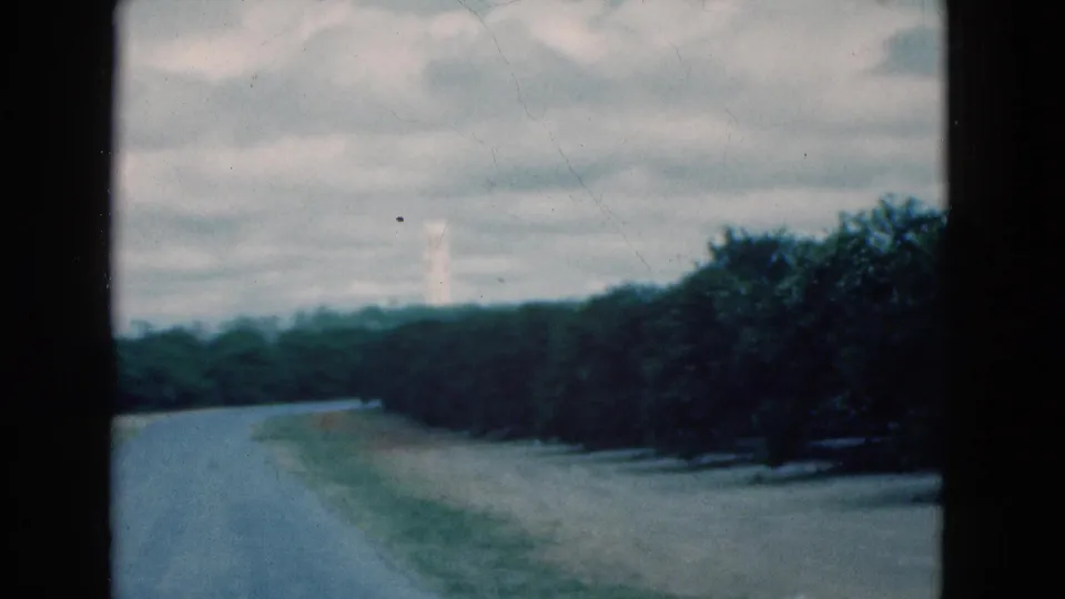 Muted 8mm frame from 1975 Florida showing a deserted road bordered by trees and a faint white tower in the distance.
