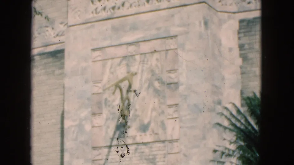 1975 Florida home-movie frame of a pale carved monument or wall panel with inscription-like relief details and nearby foliage.