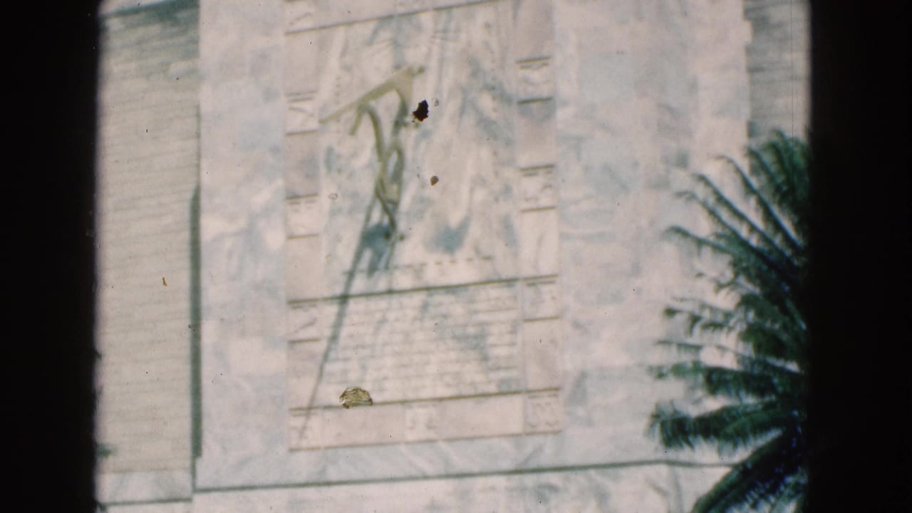 1975 Florida 8mm frame showing a pale stone monument panel with a carved relief figure, inscription area, palm shadow, and dark film border.