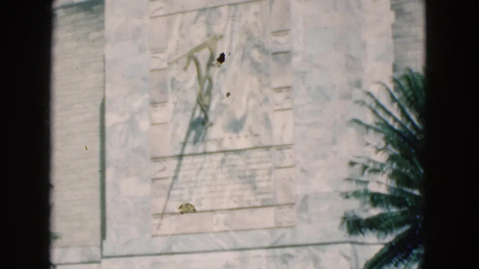 1975 Florida 8mm frame showing a pale stone monument panel with a carved relief figure, inscription area, palm shadow, and dark film border.