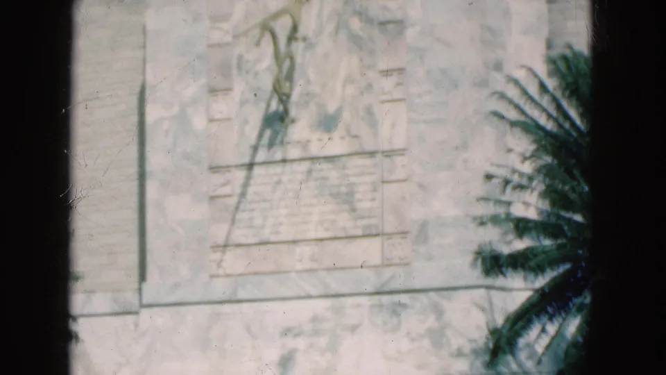 1975 Florida 8mm frame of a carved stone monument panel, inscription block, and palm frond partly crossing the right side of the view.