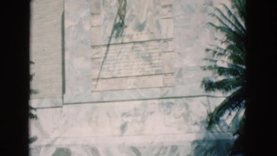 1975 Florida 8mm frame showing the lower portion of a pale stone monument panel, carved detail, inscription area, and palm foliage at the side.