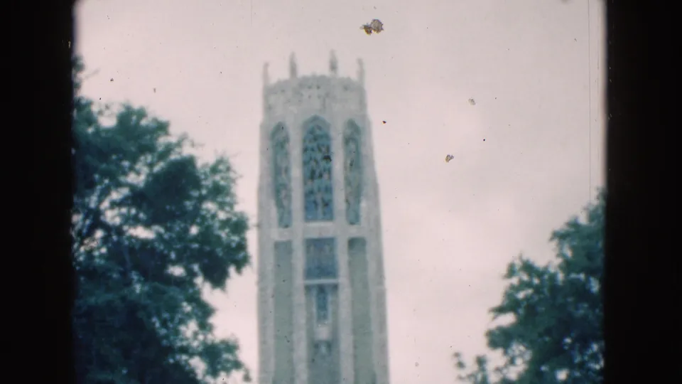 1975 Florida 8mm frame of a pale stone bell tower with arched openings, seen through trees against an overcast sky.