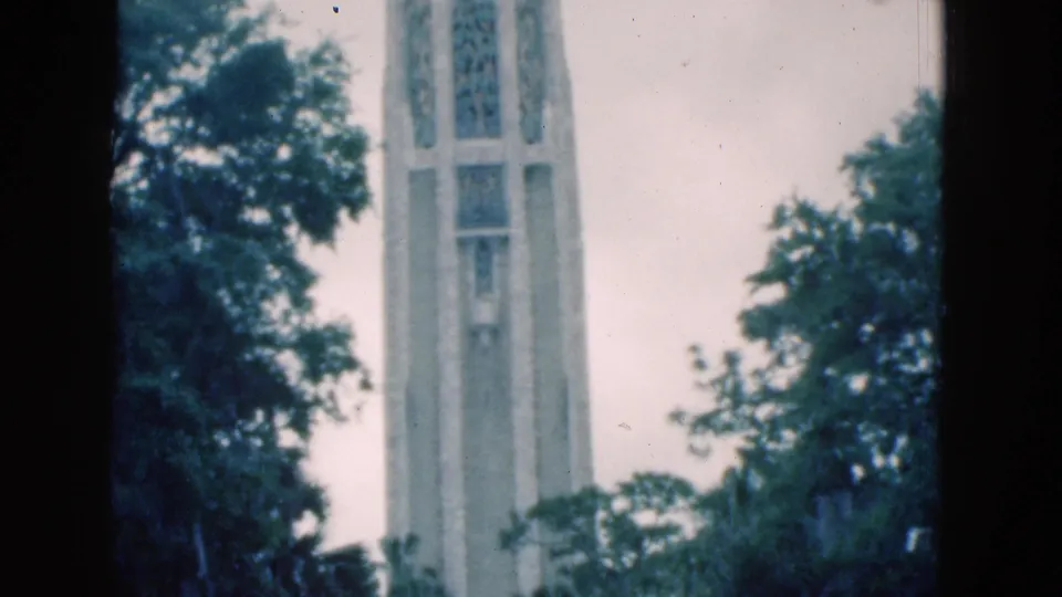Final 8mm frame of the stone bell tower, with Gothic-style openings, surrounding trees, and a pale overcast sky.