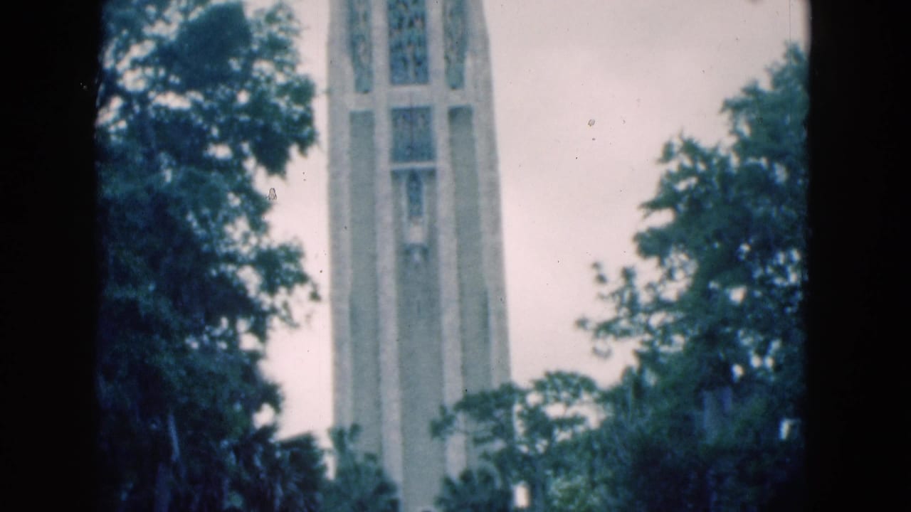 Tall pale stone tower rising behind dark green trees under a cloudy sky.