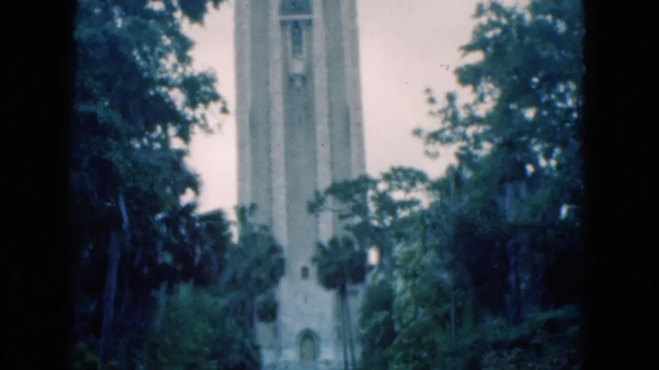 1975 Florida 8mm frame showing the tower base through trees, with small pedestrians on the grounds.