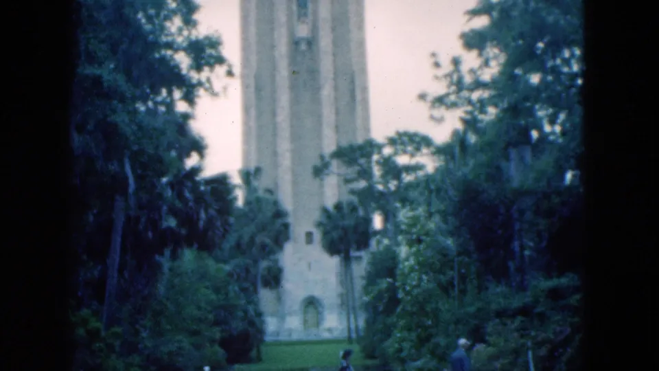 1975 Florida home-movie frame with two distant walkers crossing near a tree-lined stone bell tower.