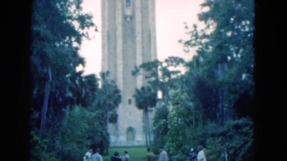 1975 Florida home-movie frame showing small visitor groups in a green park with palm trees and a tall historic stone tower behind them.