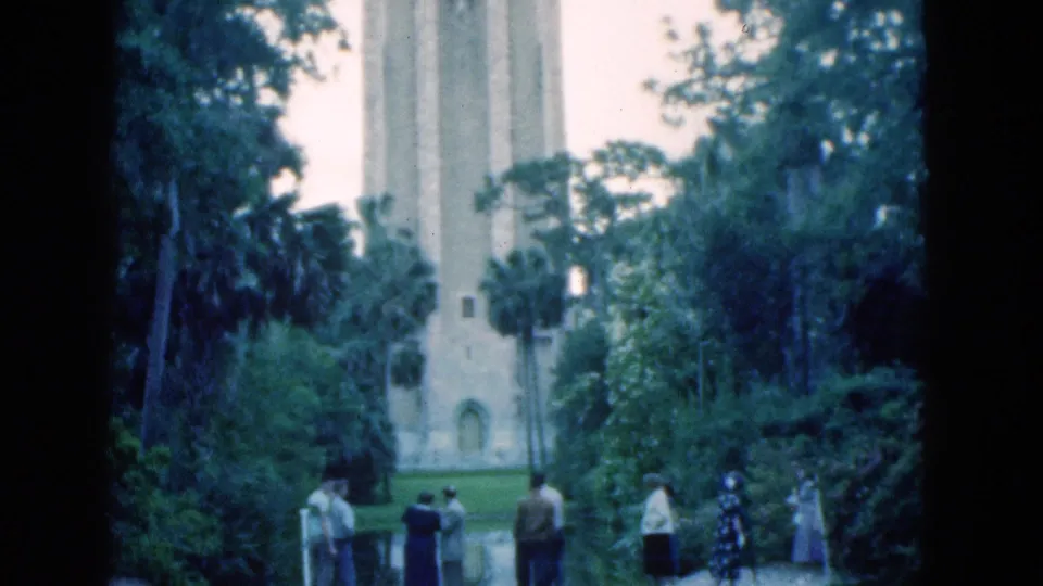 Small groups of visitors in 1970s attire stand near a curved path and water edge in a Florida park beneath a pale tower.