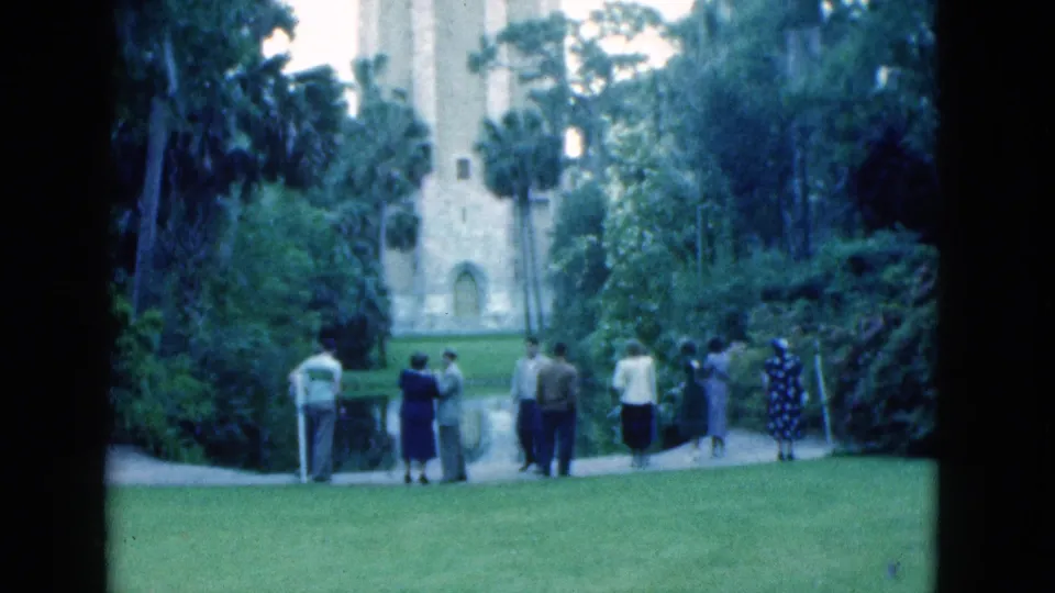 Wide 8mm frame of a Florida landmark park in 1975, with visitors on the lawn and path below a tall stone tower and palms.