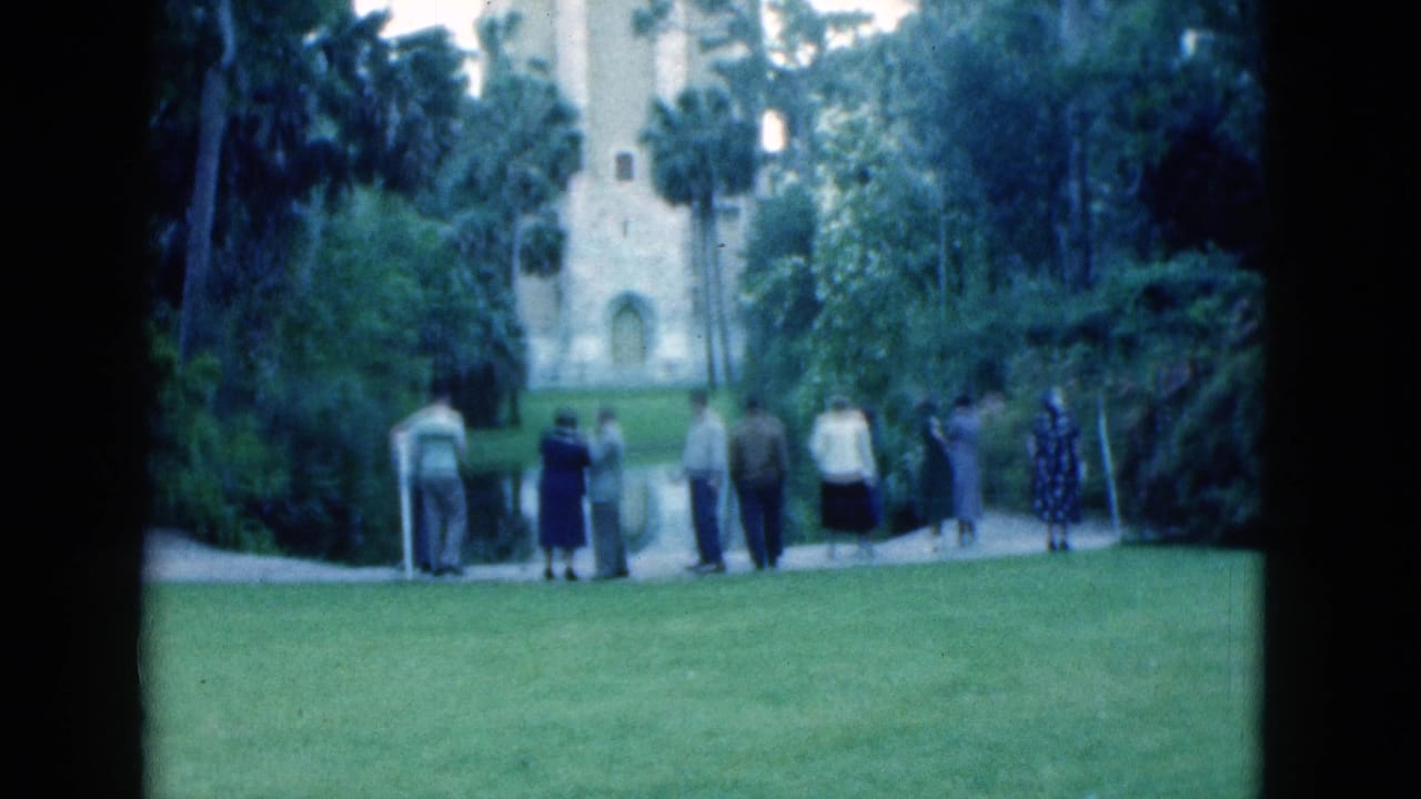 Adults stand on a lawn near a pale historic-looking building and palm trees.