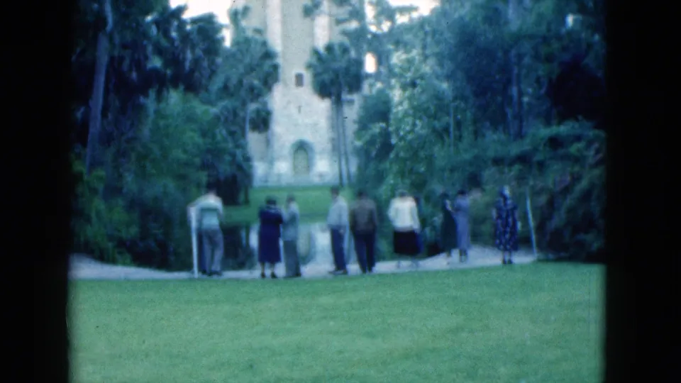 1975 Florida 8mm frame showing adults gathered on grass near a pale stone building, path, palms, and dense foliage.