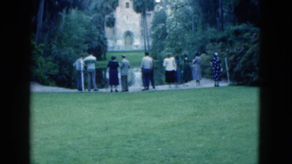 1975 Florida home-movie frame with adults standing near a path, facing a pale historic-looking building amid tropical landscaping.