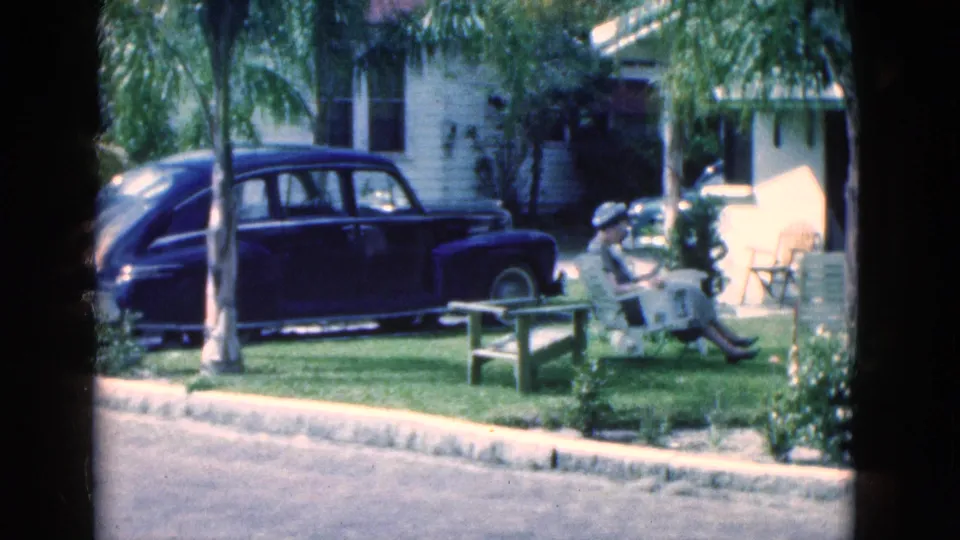 In a Florida residential yard, a woman in a sunhat sits reading near a dark mid-century sedan, palm trees, and a light-colored house.