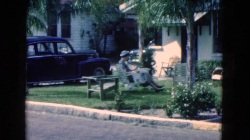 The 1975 Florida home movie shows a woman seated in a yard reading, with a dark sedan, palm trees, and a residential exterior nearby.