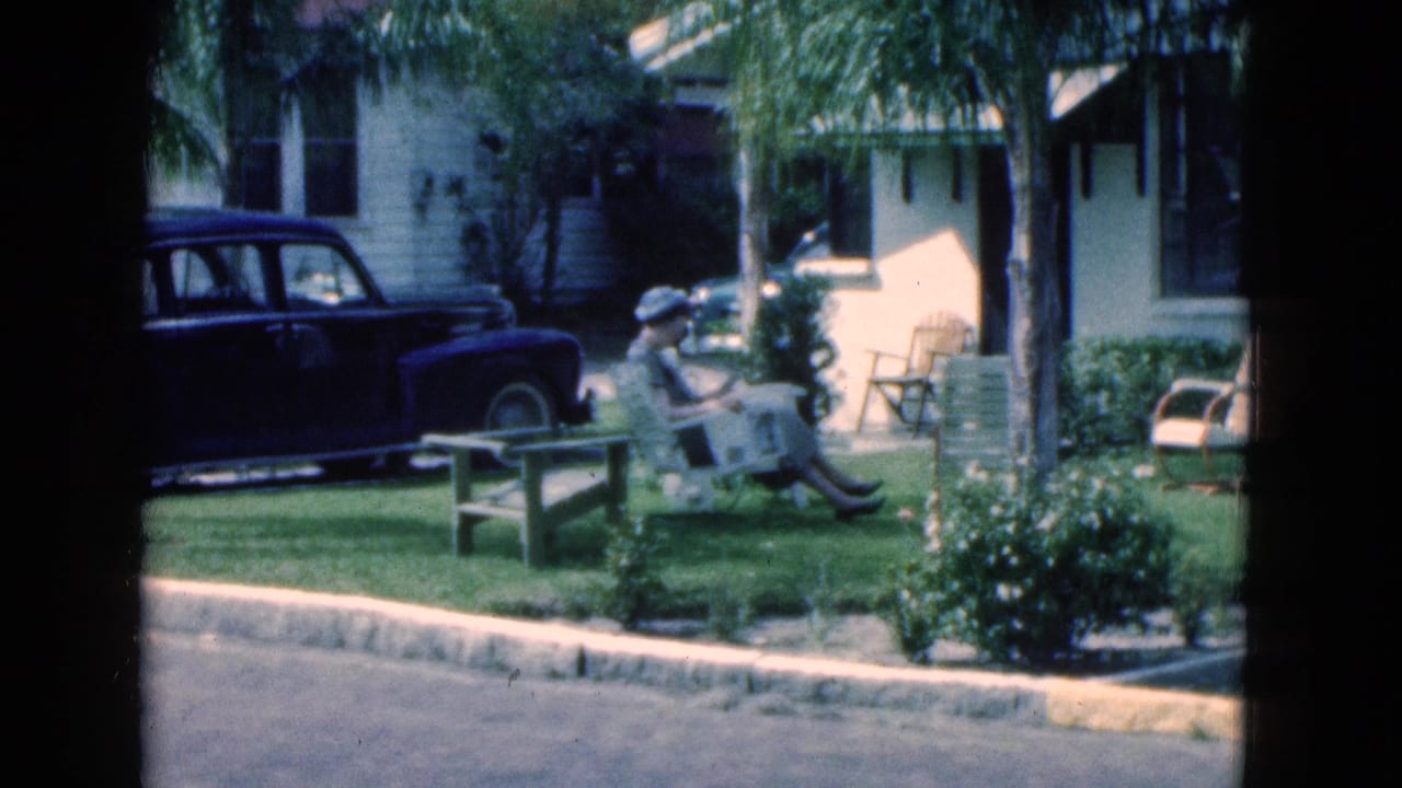 In 1975 Florida home-movie footage, a woman sits on a bench reading in a sunny yard beside a dark sedan and stucco house.