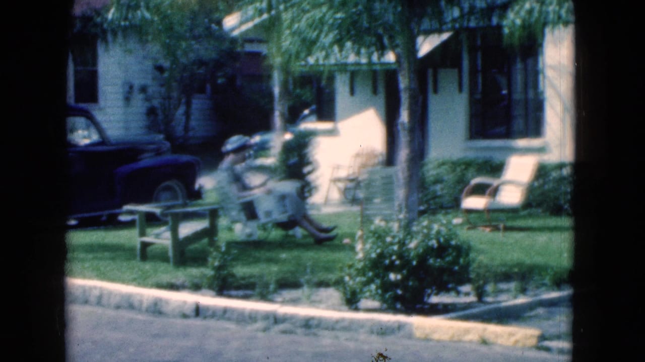 The woman remains seated reading in a landscaped Florida yard, with chairs, shrubs, palms, and the house exterior visible.