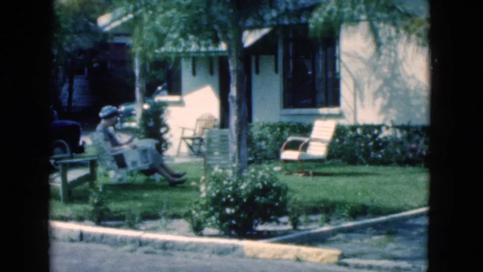 A slightly shifted 8mm frame shows the woman reading on the bench with the lawn, shrubs, chairs, and stucco house behind her.